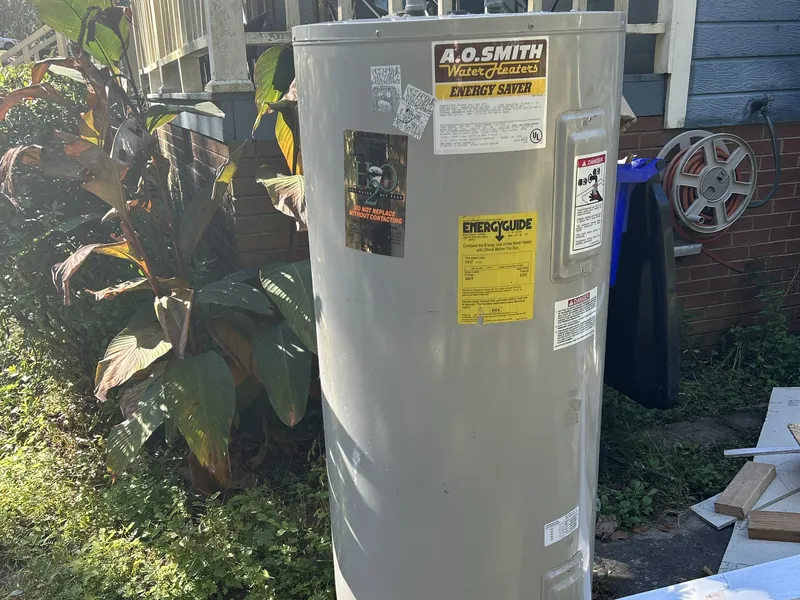 An old electric water heater standing beside a house, ready to be hauled away