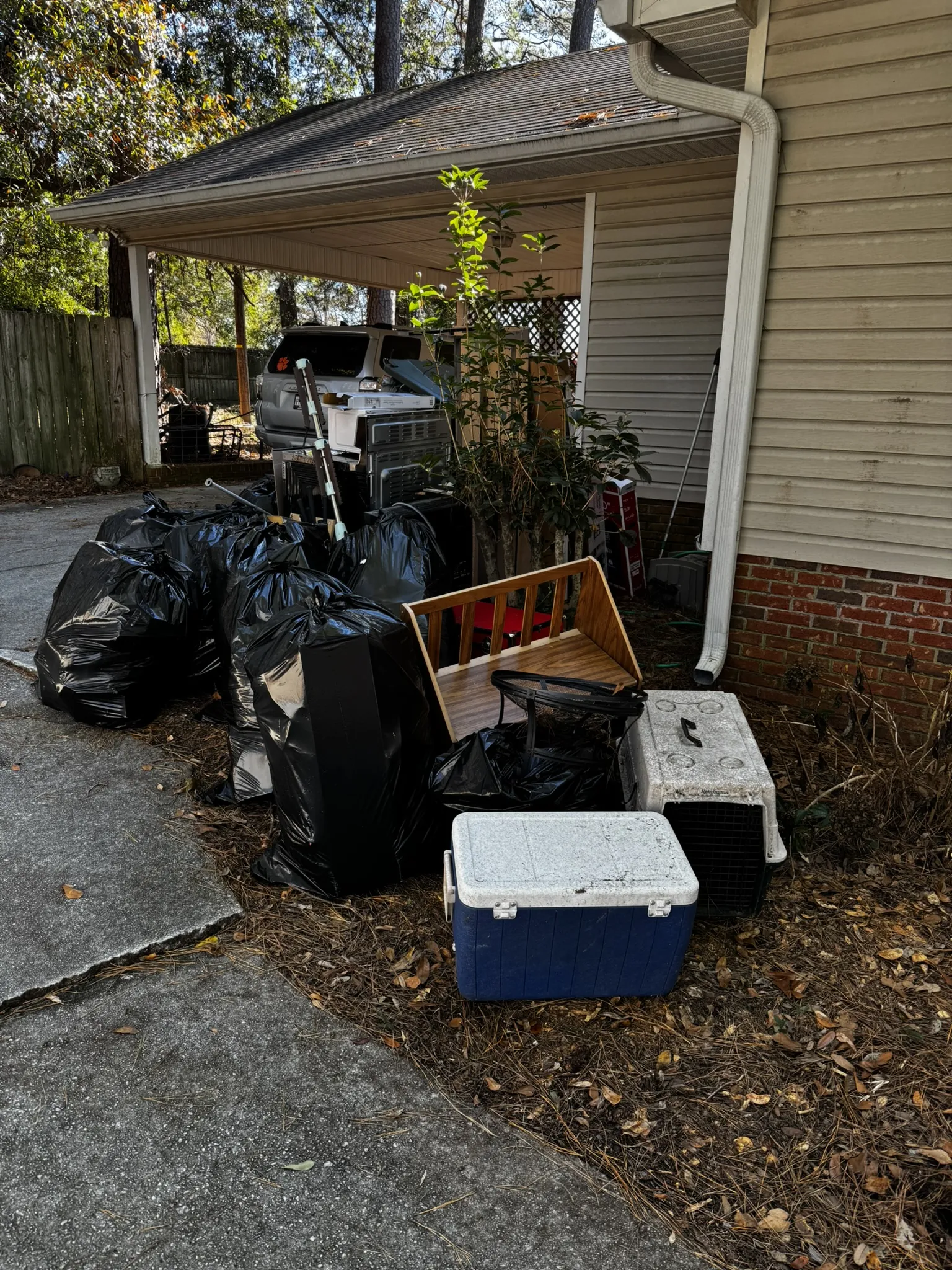 Trash bags, a cooler, a pet crate and a wooden crate by a carport