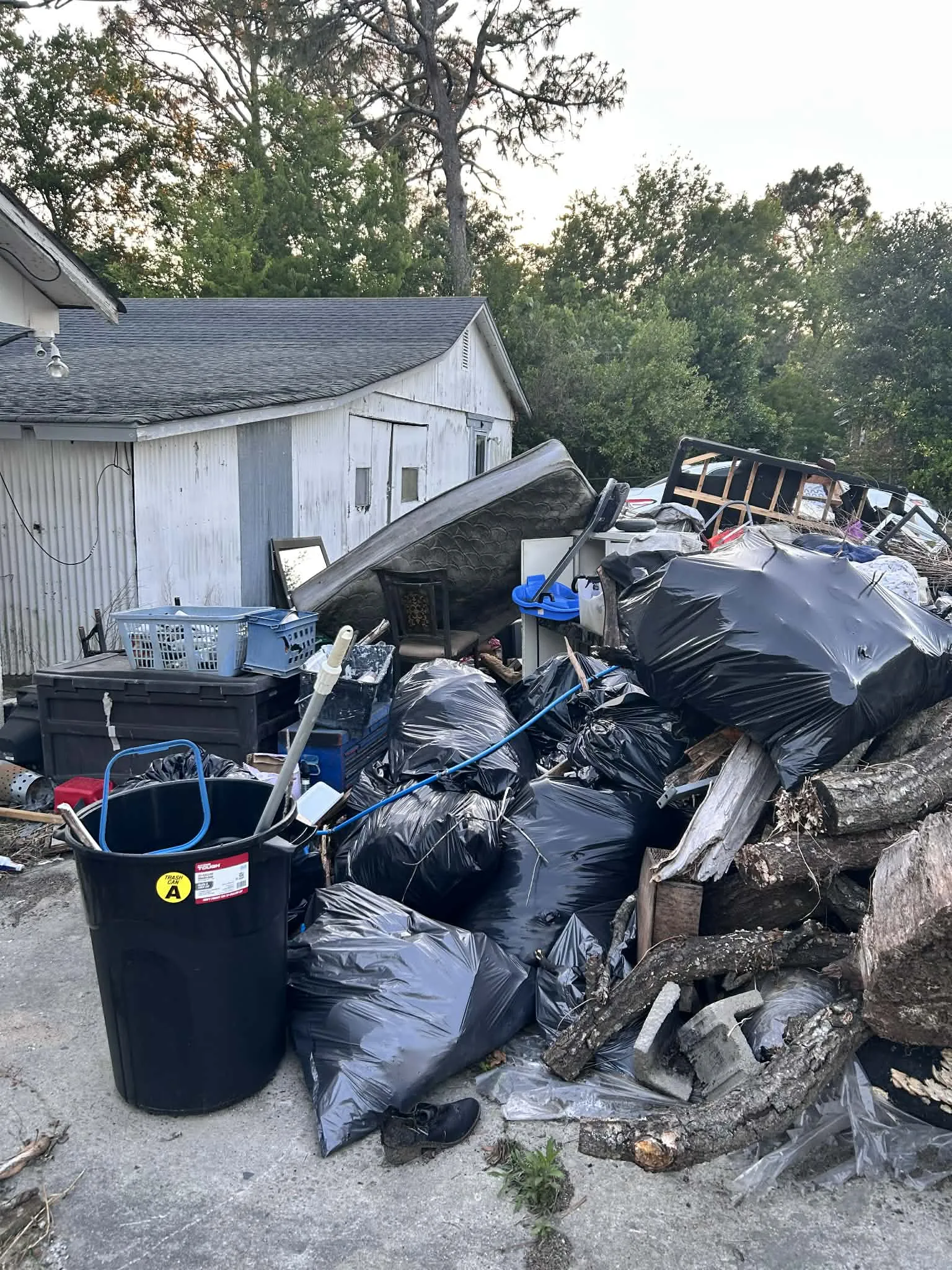 A large pile of trash bags, a mattress, bins and logs beside a shed