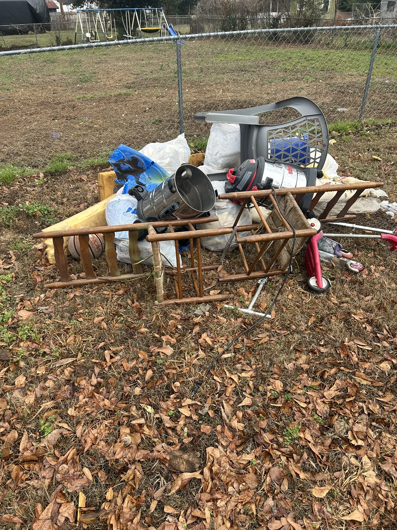 A pile of household items and a wooden ladder in a fenced yard, before pickup