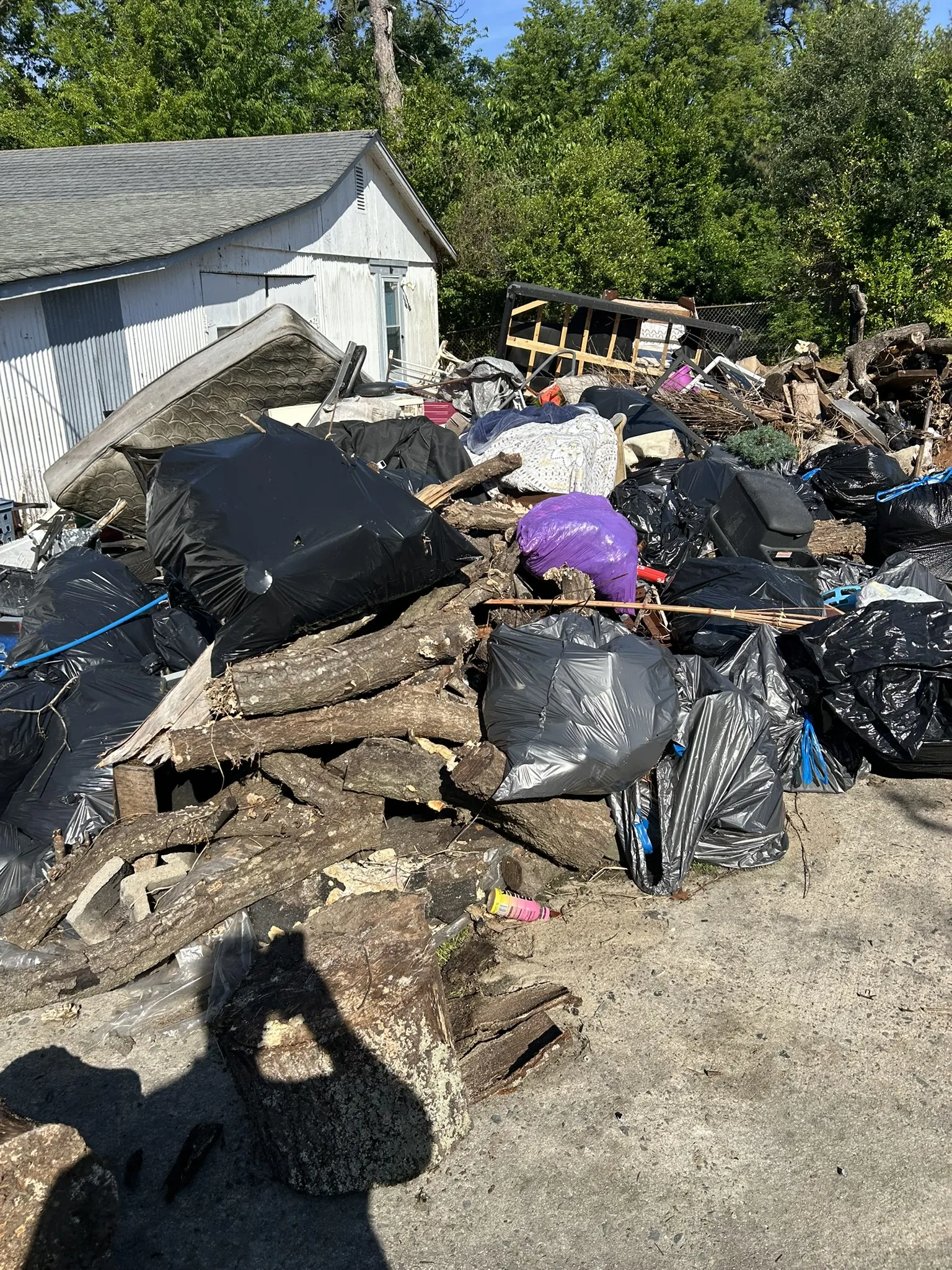 A large pile of trash bags, logs and a mattress beside a shed