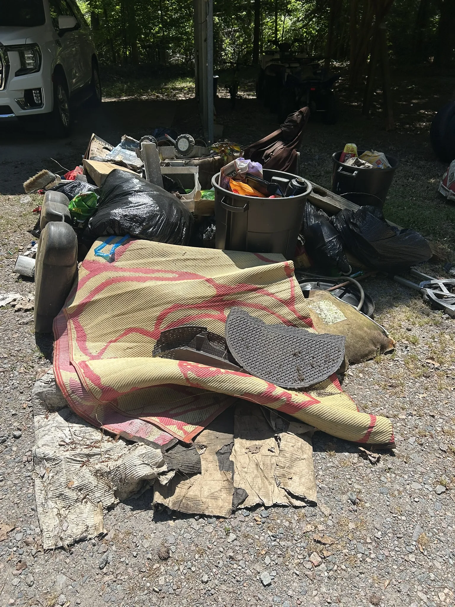 A rug, trash bags, bins and old items piled under a carport